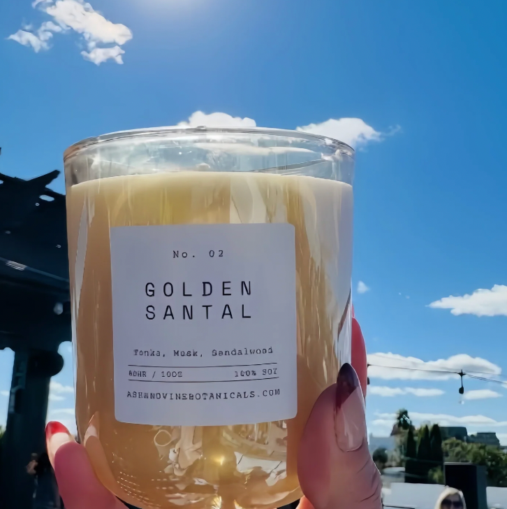 Candle labeled 'Golden Santal' held against a blue sky with clouds