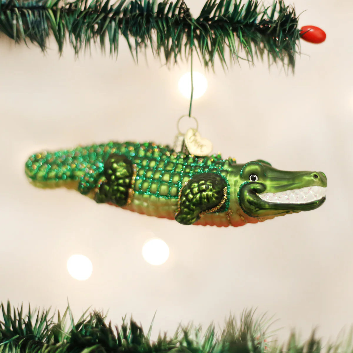 A green alligator-shaped ornament hanging from a Christmas tree with a crocodile-like face and a red berry hanging from its mouth.