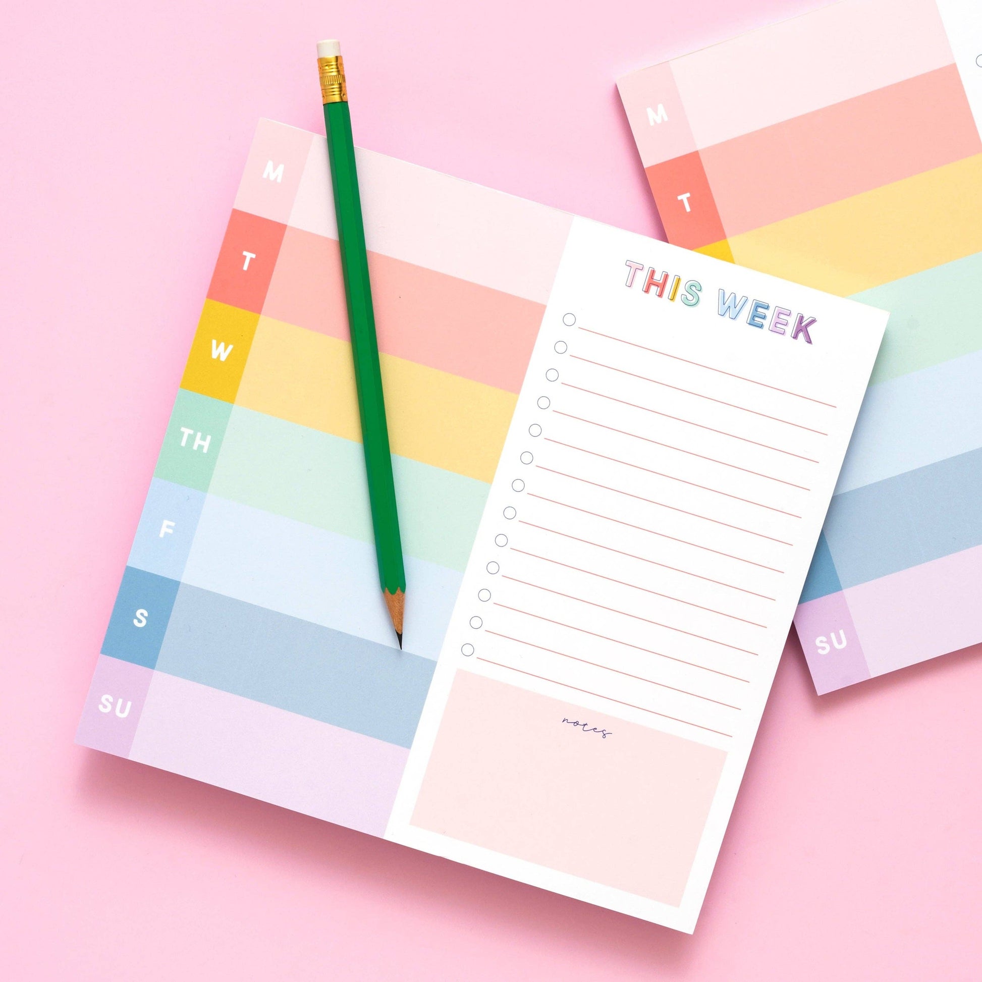 Colorful weekly planner notepad with a green pencil on a pink background