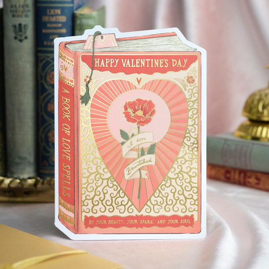 Valentine's Day card designed like a book with a heart and rose illustration, placed on a surface with books in the background.