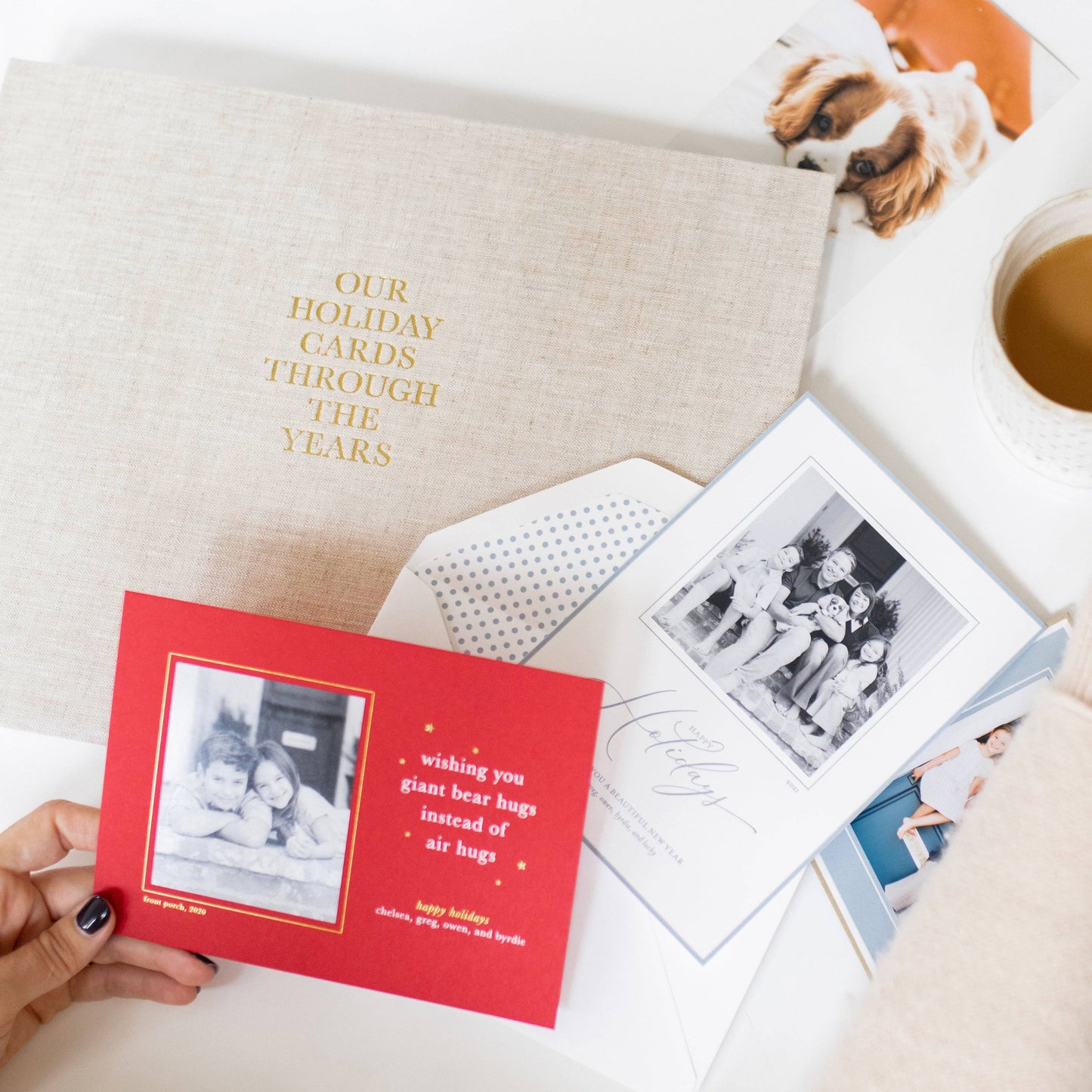 Person holding a red holiday card with a photo of two children, surrounded by other cards and a cup of coffee.